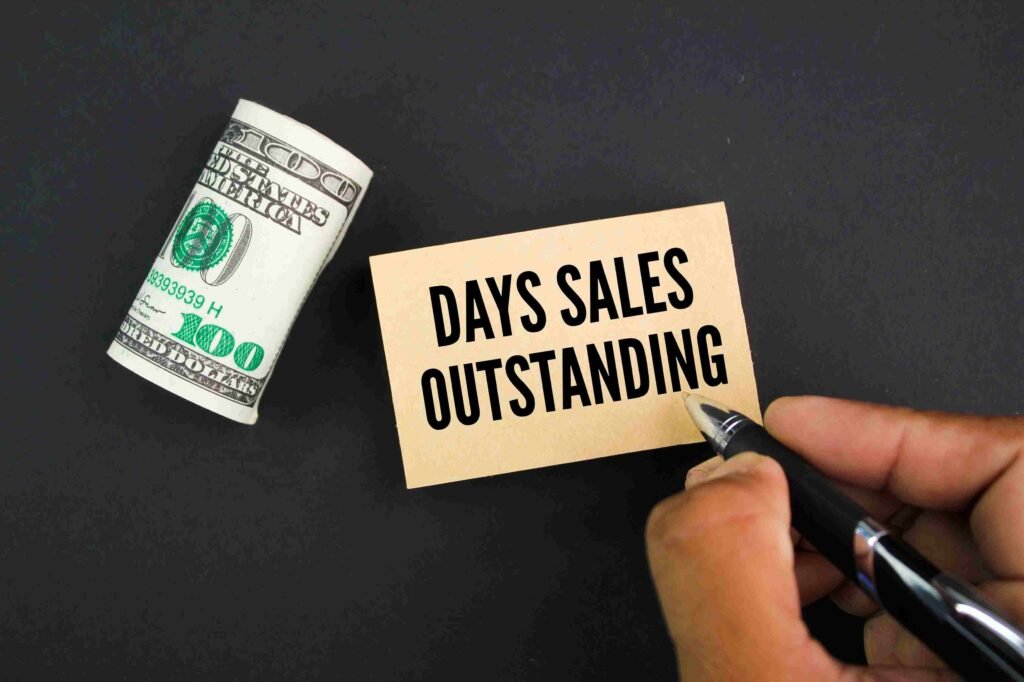 Hand holding a pen writing Days Sales Outstanding on a card next to a roll of US dollar bills