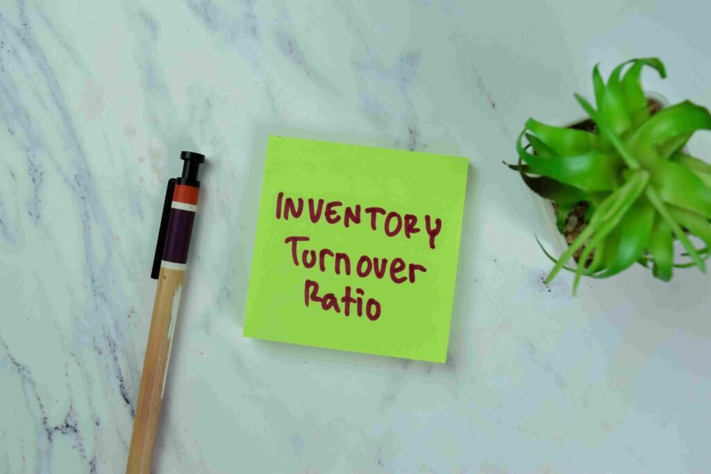 Sticky note labeled inventory turnover ratio placed beside a pen and small plant on a desk